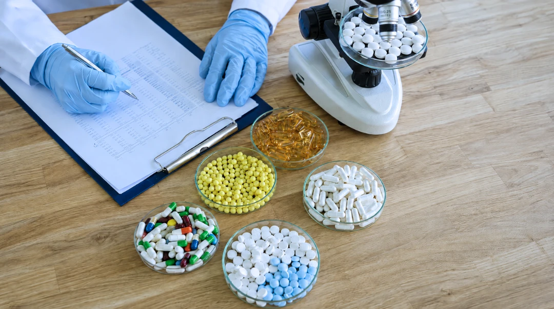 Pharmaceutical scientist recording data beside capsules, tablets and pellets in petri dishes with a microscope